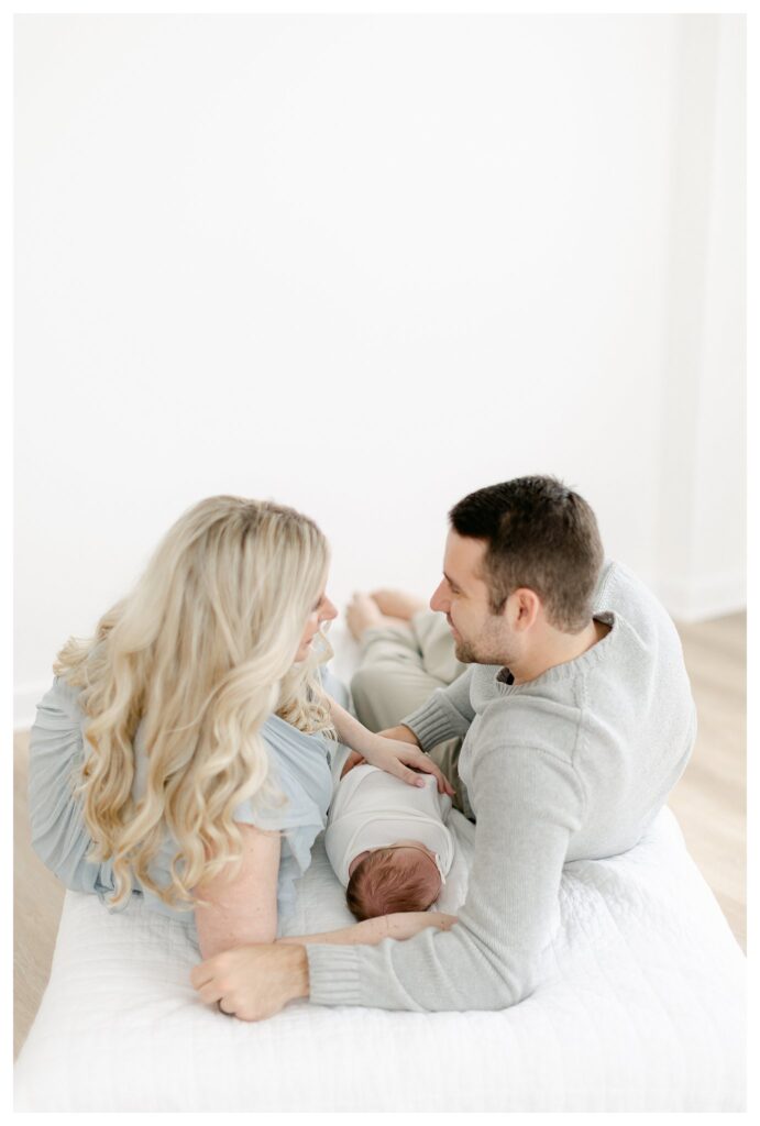 Mom and dad laying on a white bed with their babygirl in a natural light studio captured by Philadelphia Newborn Photographer Tara Federico