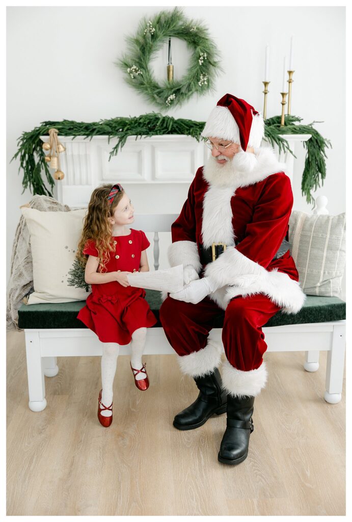 Santa sitting on a bench with a sweet little girl who is showing him her Christmas Wish list during South Jersey Santa Pictures with Tara Federico Photography