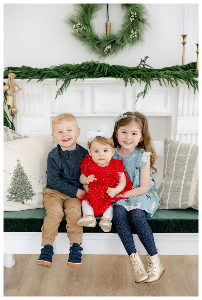 A little boy, a little girl and their baby sister sitting on a bench in front of a fireplace during their South Jersey Santa Pictures with Tara Federico Photography