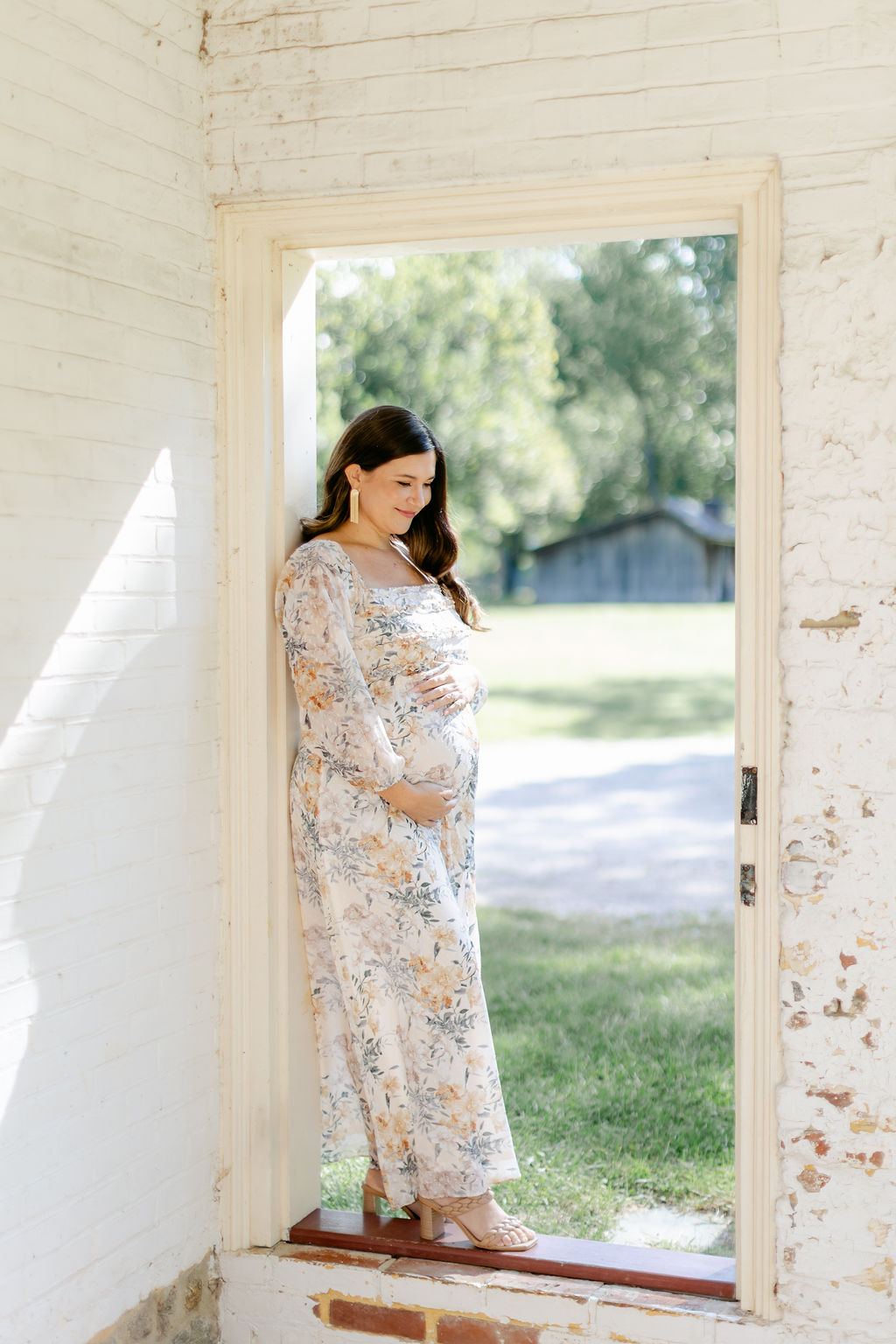 A mother to be in a white floral printed dress stands ina. doorframe of an old building in a park