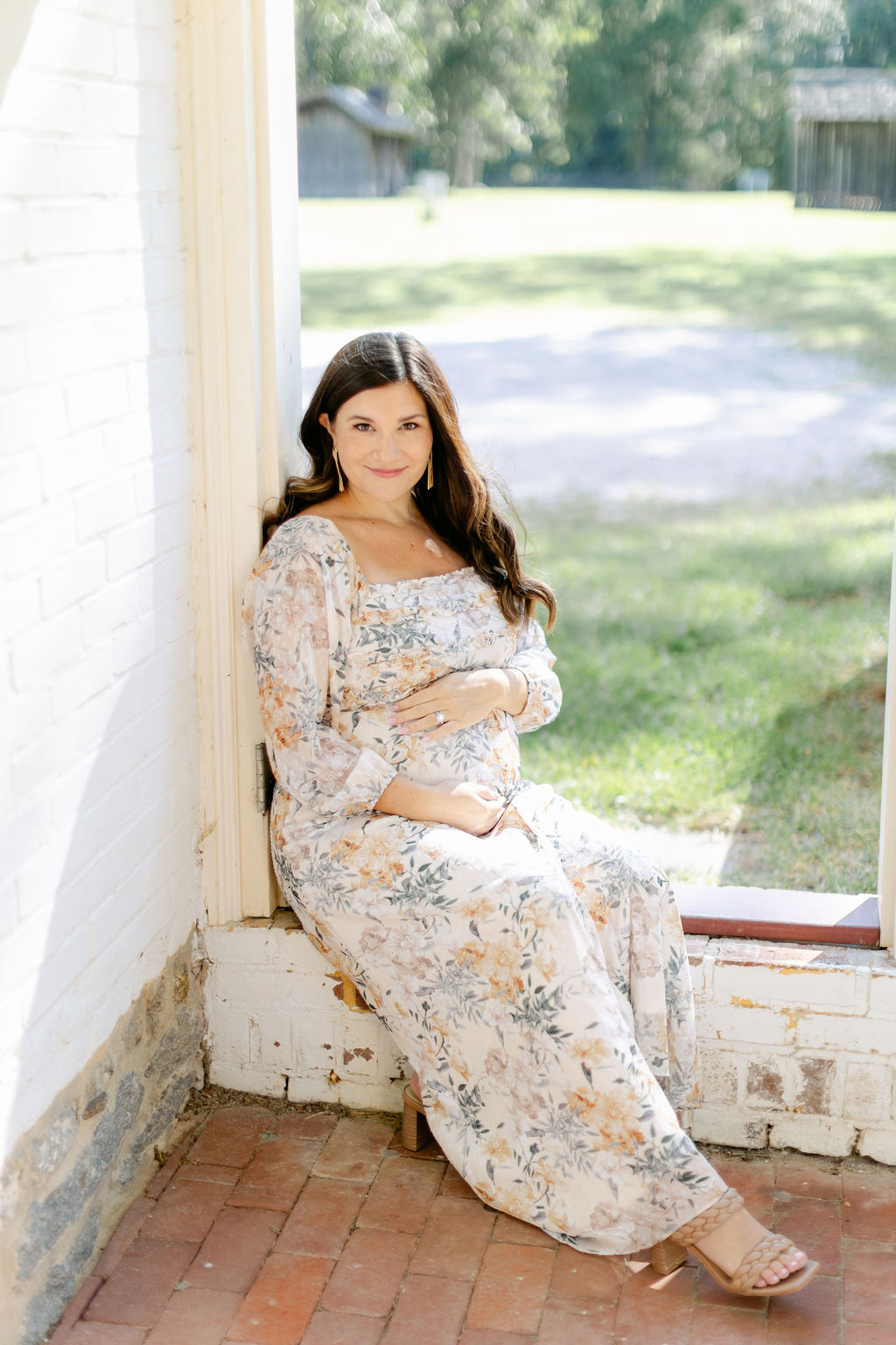 A happy expecting mother in a floral dress sits on a brick porch in a park before visiting baby shower venues charleston sc