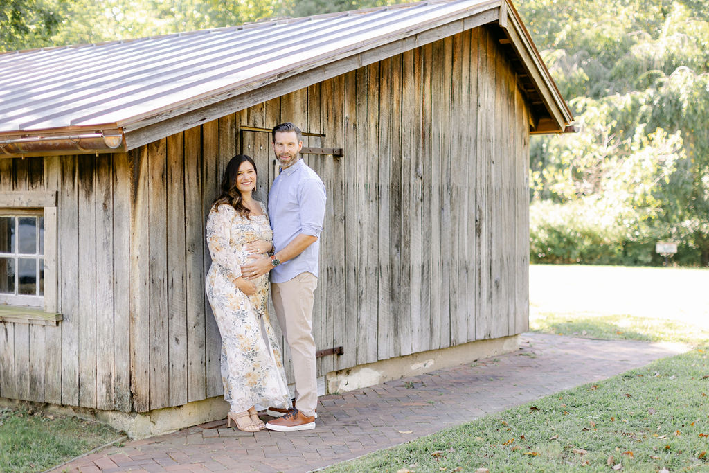 A mother to be leans against an old wooden building in a park with dad holding the bump before visiting baby shower venues charleston sc