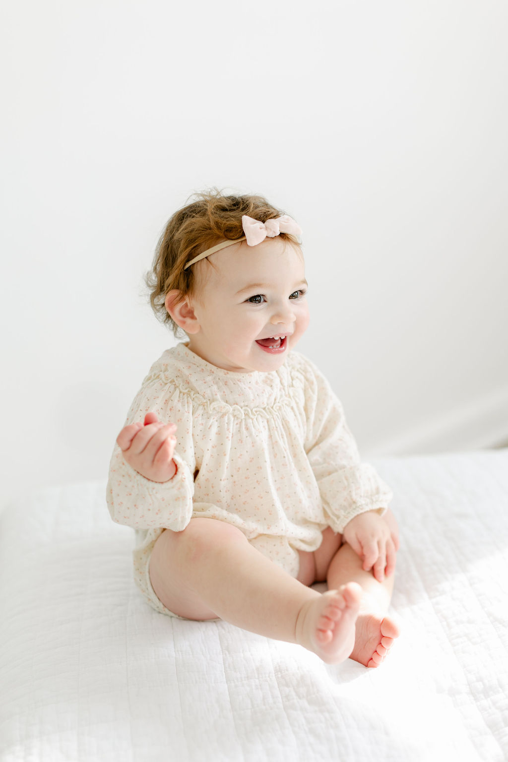 A happy infant girl sits on a bed smiling and laughing in a pink bow