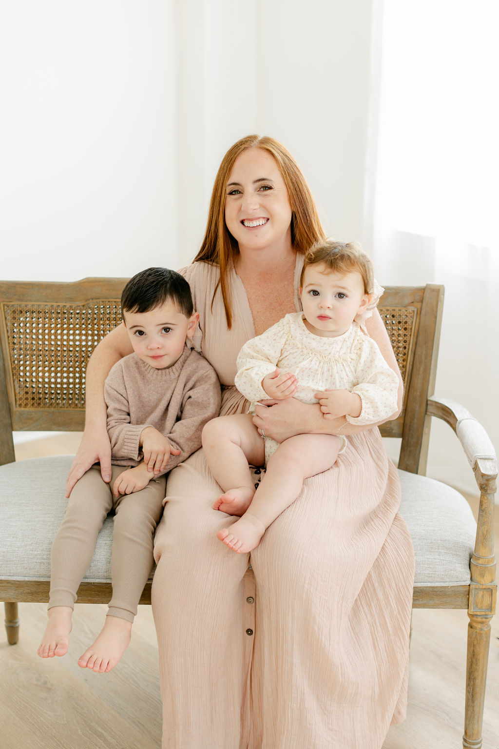 A happy mother sits on a bench in a studio with her toddler in her lap and older toddler son at her side before visiting baby stores charleston sc