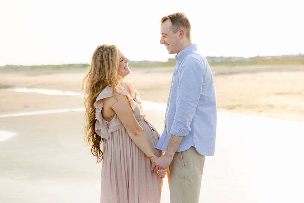 Expecting parents stand on a beach holding hands and smiling at each other on one of the beaches near charleston sc at sunset