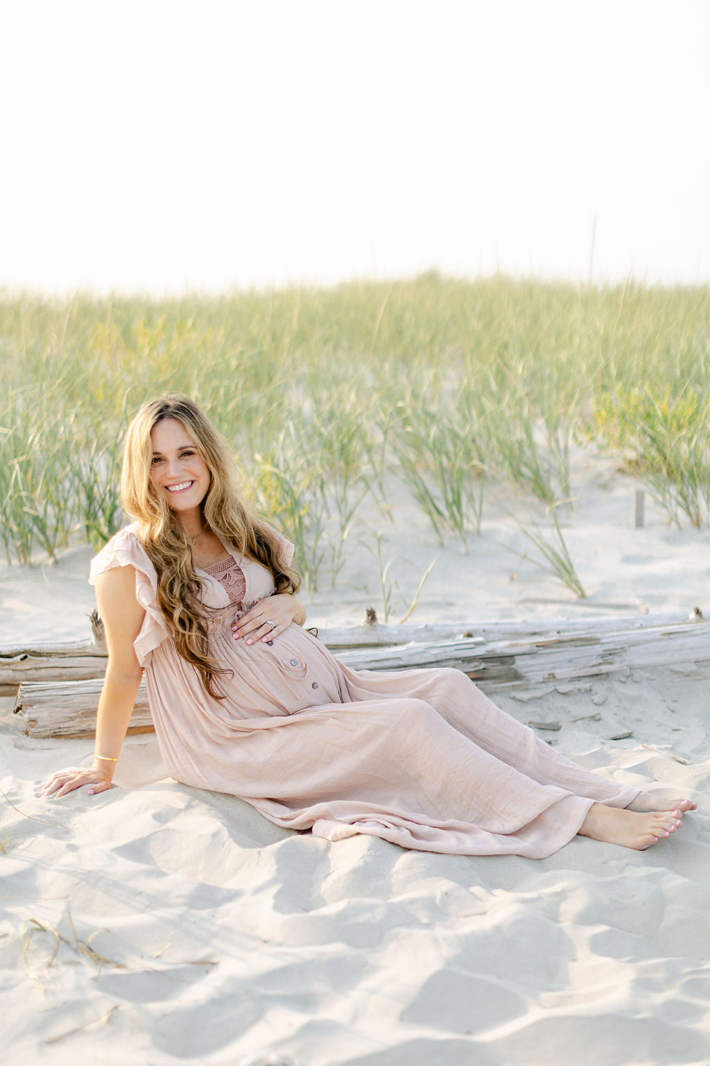 A mom to be in a pink dress sits on a beach dune at sunset holding her bump