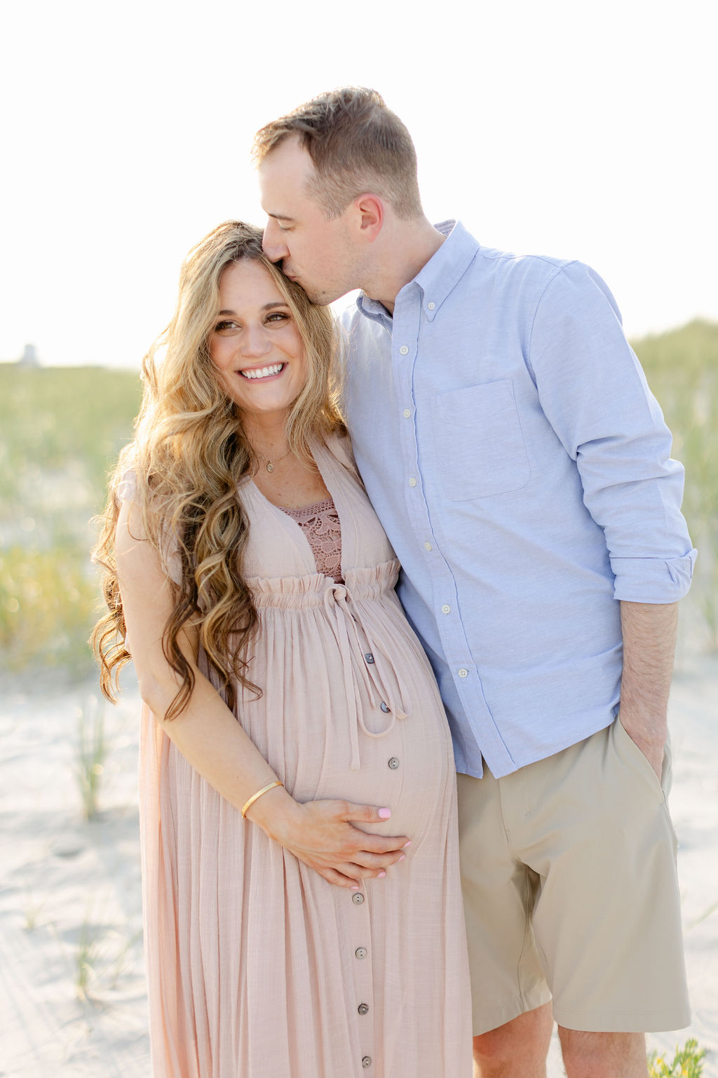 A husband in a blue shirt kisses the head of his pregnant wife while standing on a beach at sunset after visiting birth center charleston sc