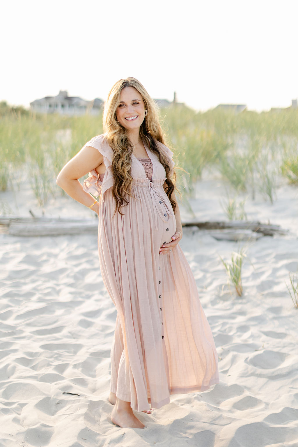 A mother to be in a pink maternity dress stands on a beach at sunset holding her bump after visiting a birth center charleston sc