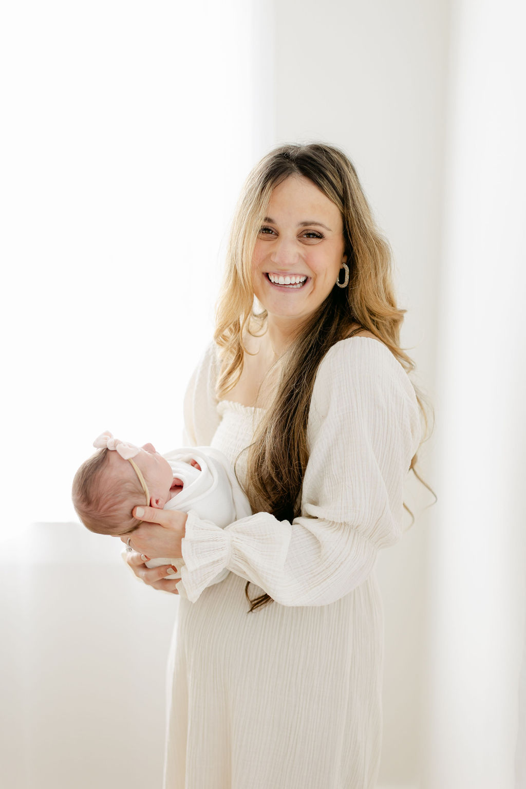 A happy new mother in a white dress stands in a studio holding her sleeping newborn baby girl in a pink bow and swaddle after meeting with charleston sc pelvic floor therapists