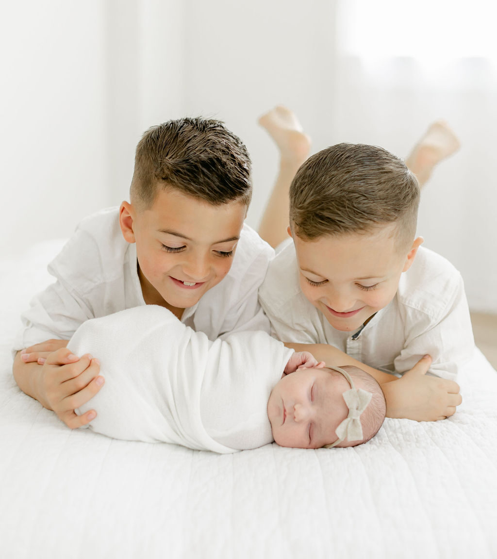 Two young boys lay on a bed smiling down at their newborn baby sister sleeping in front of them taken by a mount pleasant family photographer
