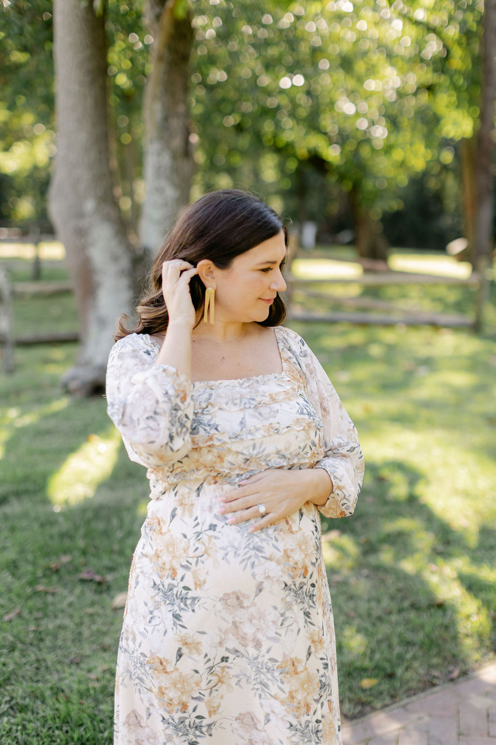 A mother to be in a yellow floral dress brushes her hair behind her ear while standing in a park after visiting yoga studios charleston sc