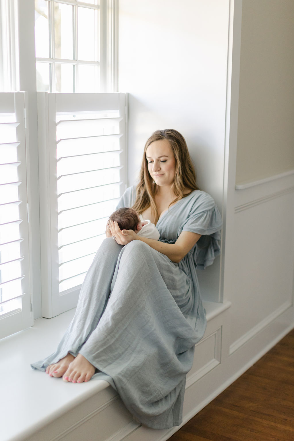 A happy new mom sits across a window sill in a studio cradling her newborn baby in her lap with help from charleston sc newborn photography