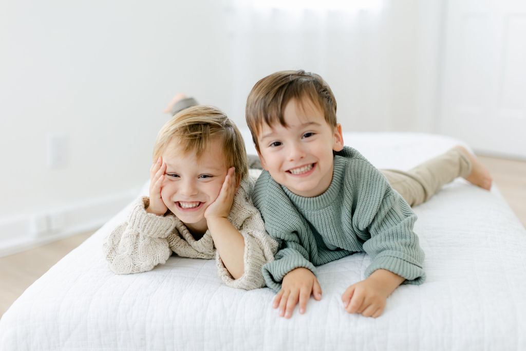Two toddler brothers lay on a bed together in knit sweaters