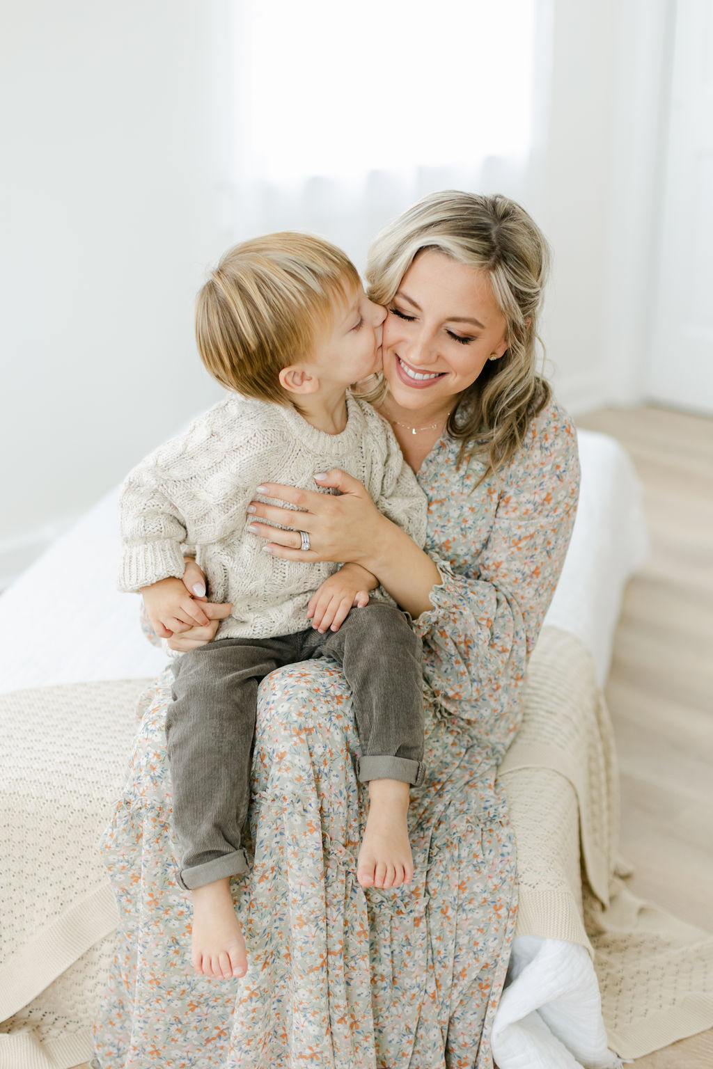 A happy mother sits on a bed in a studio with her toddler son kissing her cheek while sitting in her lap before visiting family friendly restaurants charleston sc