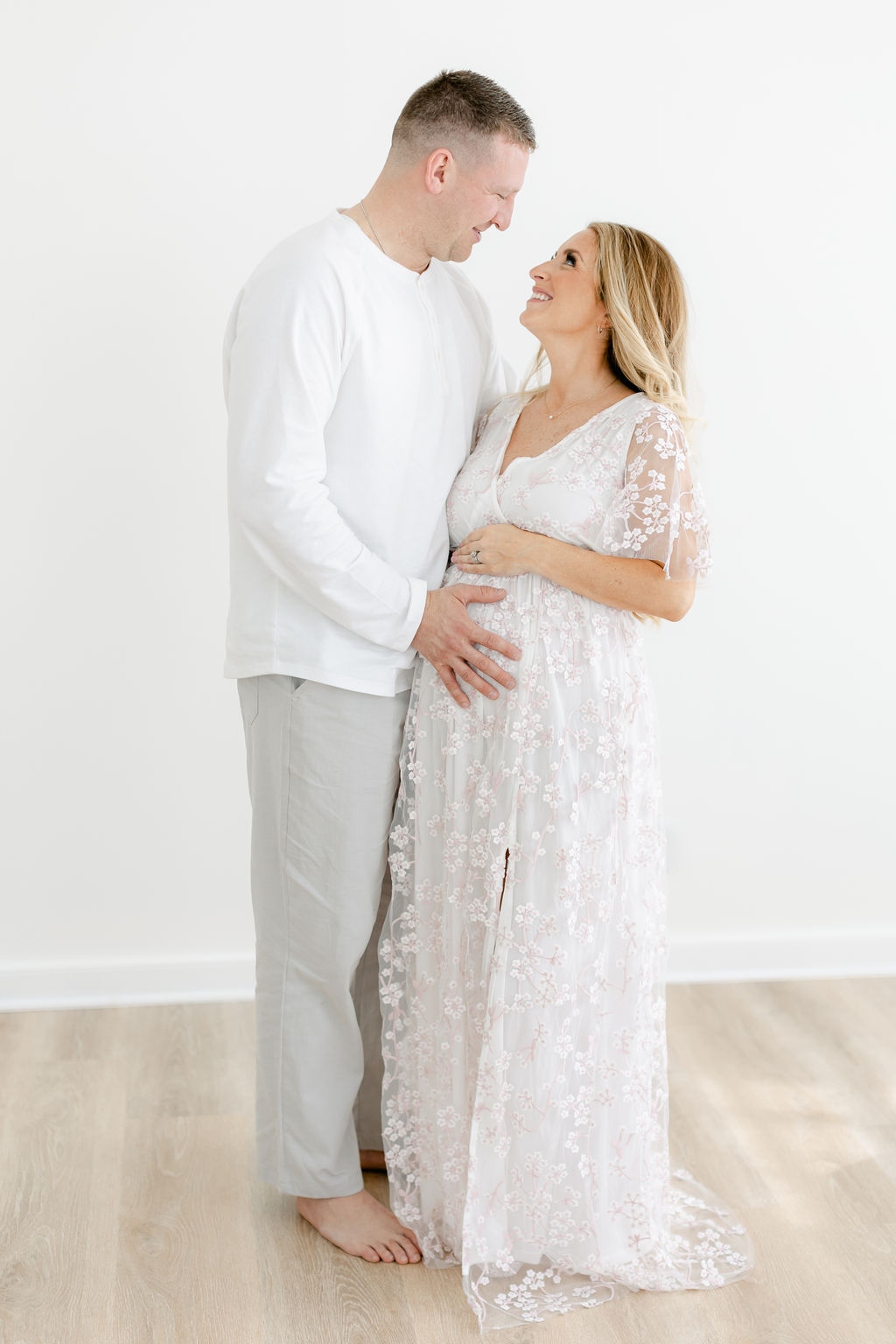 Happy expecting parents stand in a studio smiling at each other and holding the bump