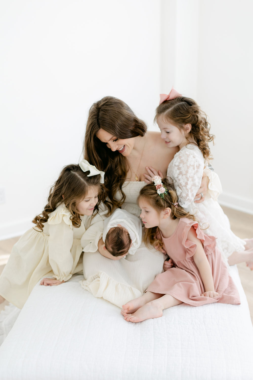 A happy mother shows her three toddler daughters their sleeping newborn baby sister on a bed in a studio