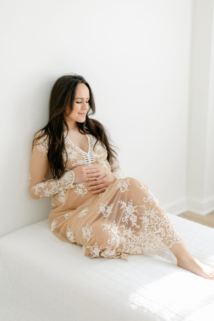 A mother to be smiles while sitting on a bed in a studio with hands on the bump in a lace maternity gown before some prenatal yoga charleston sc