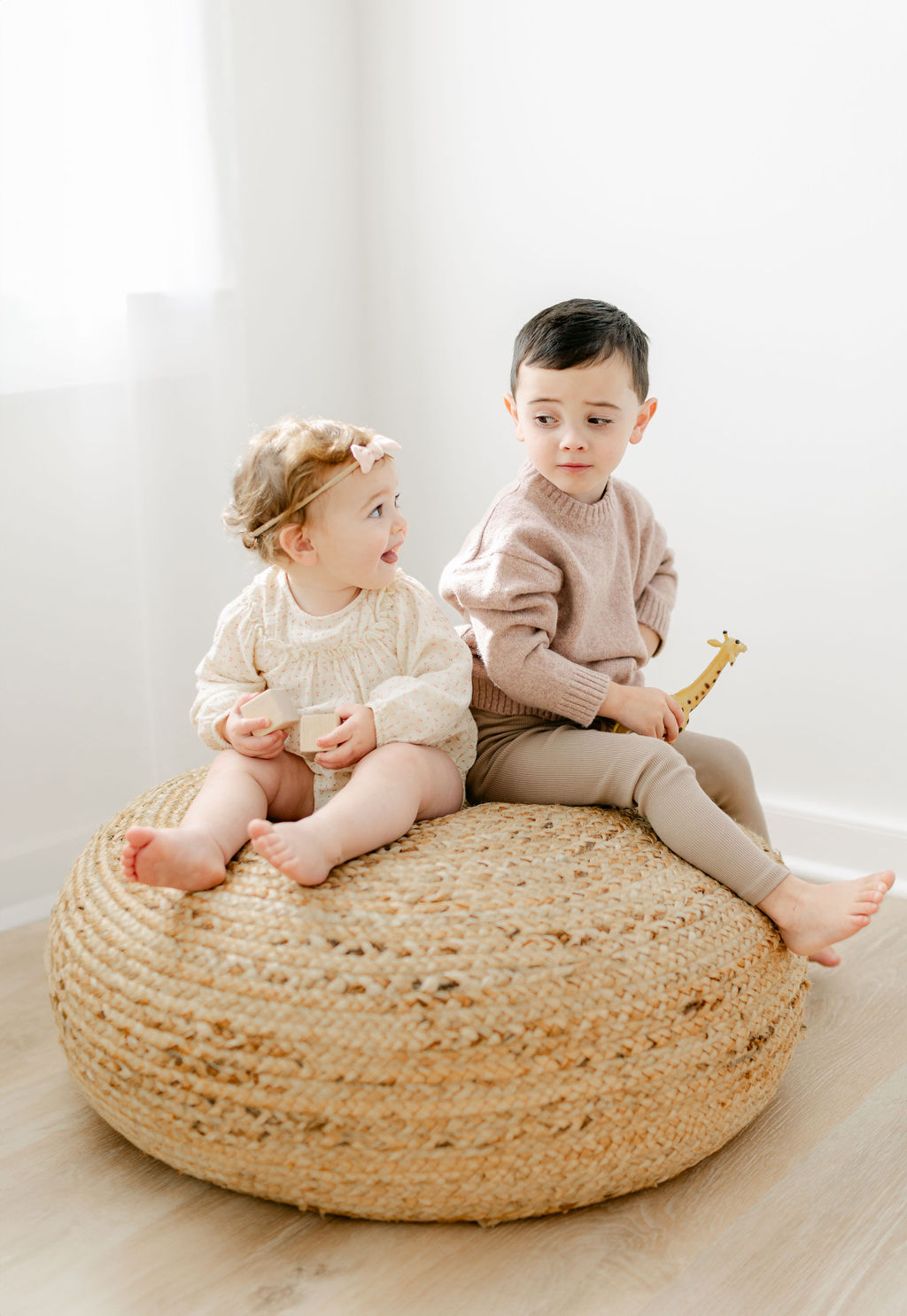 A young toddler girl and her slightly older brother sit on a woven bean bag stool in a studio while looking at each other before visiting toy store charleston sc