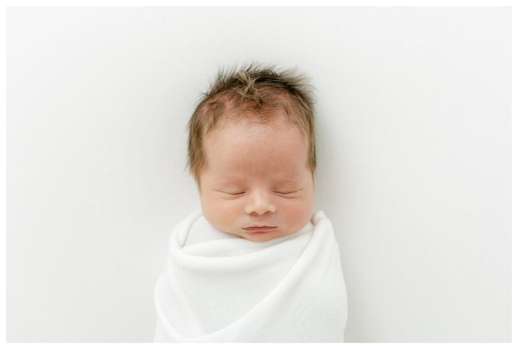 Newborn baby boy swaddled in a white blanket during a Charleston newborn session prep, peacefully sleeping in a studio setting.