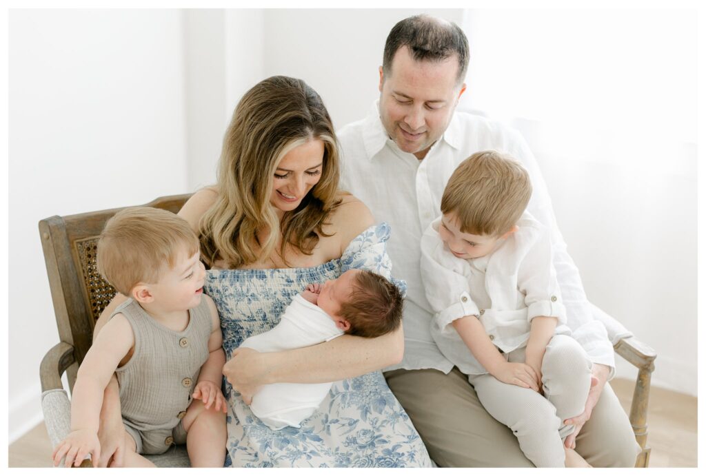 Candid moment of mom and dad with their three sons, including their newborn baby wrapped in white, and two toddler boys in neutral white and tan outfits during a Charleston newborn session prep.