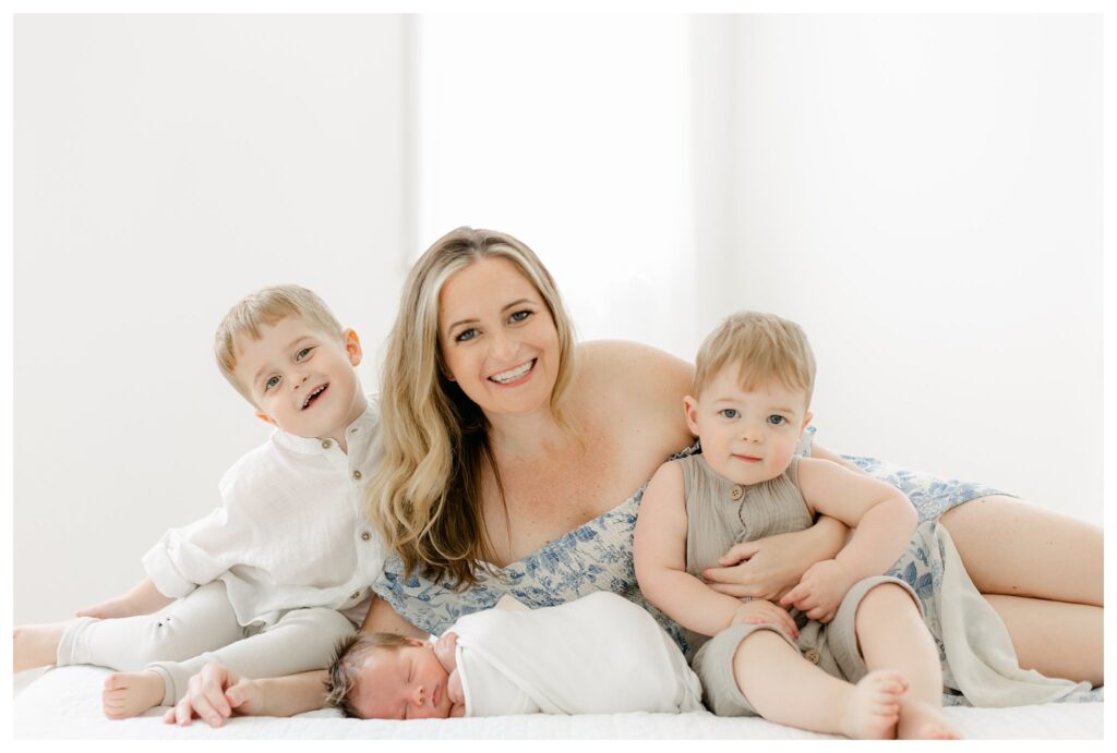 Mom with her two toddler boys and newborn baby boy wrapped in a white swaddle during a Charleston newborn session prep, with mom wearing a white and blue floral dress