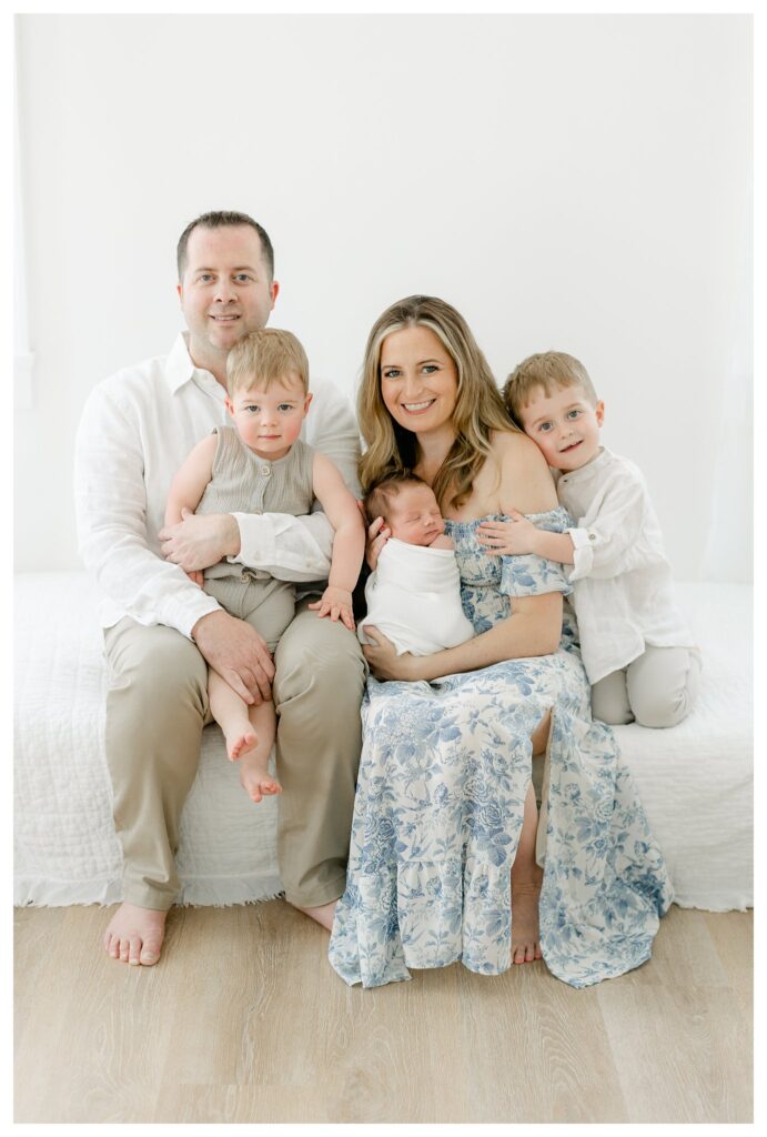 Family portrait from a Charleston newborn session prep featuring mom in a white and blue floral off-the-shoulder dress, dad, and their three boys, including the newborn baby swaddled in a white blanket.