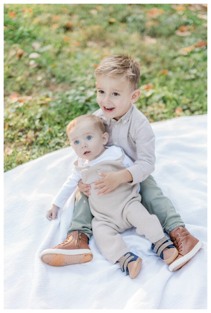 Fall family portraits in Charleston with a sweet moment between a toddler boy and his infant brother sitting on a blanket in a park.